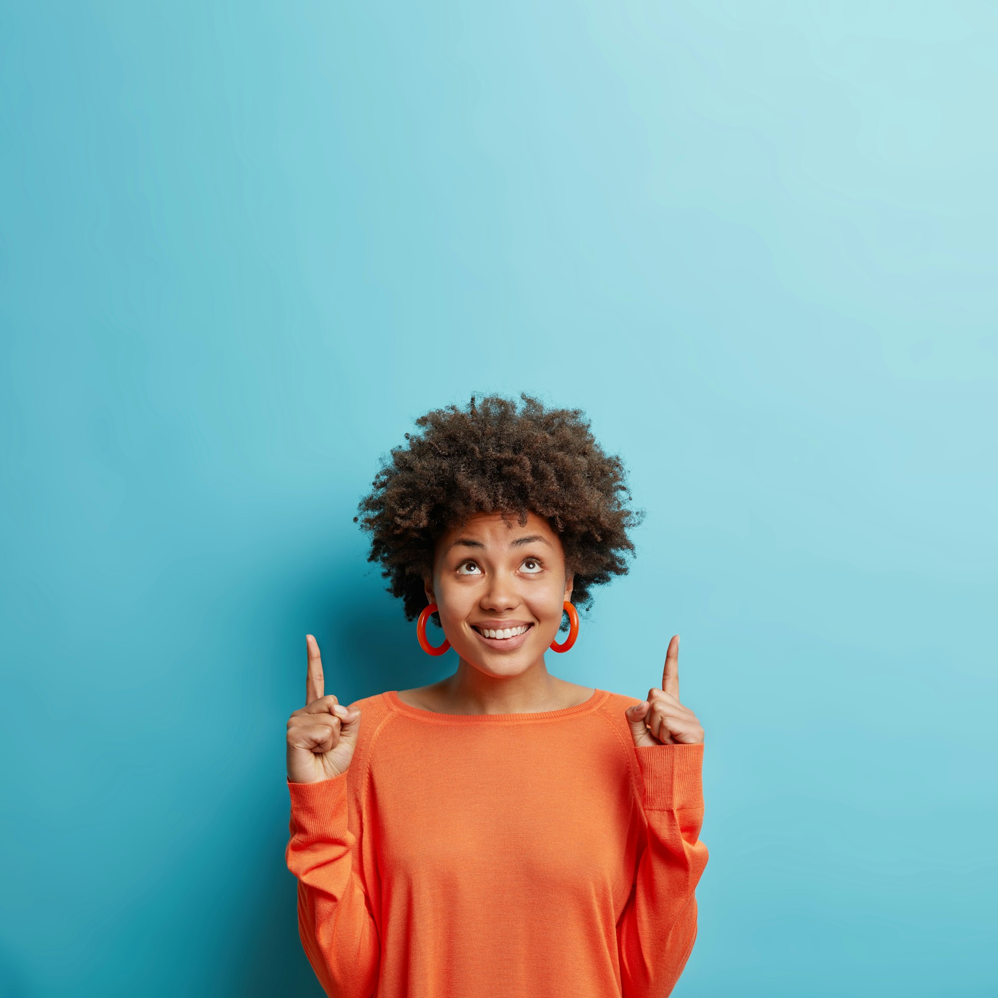 Cute young female with Afro hair points above recommends to click link shows logo and smiles broadly